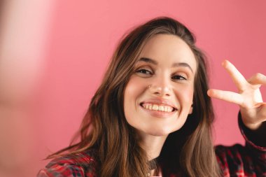 Güzel esmer bir kadının selfie çekerken, barış işareti ile poz verirken, mutlu gülümserken, fotoğraf çekerken, pembe arka planda doğrudan kameraya poz verirken..