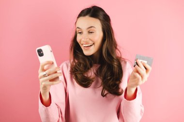 İnternetten alışveriş. Gülümseyen esmer kadın akıllı telefon uygulamasında kredi kartıyla ödeme yapıyor cep telefonu ve banka kartıyla pembe arka planda duruyor. Mutlu çığlık, vay canına, duyguları kazan..