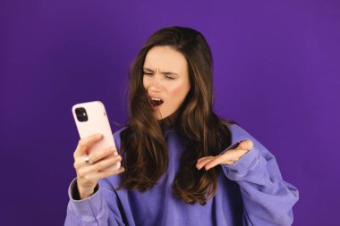 Mor arka planda izole edilmiş cep telefonu kullanan genç ve güzel bir kadın. Bağıran mutsuz kadın cep telefonunu tutuyor. Hayal kırıklığına uğramış üzgün bayan, haberleri dehşete düşürdü. Bu da ne? Olamaz.
