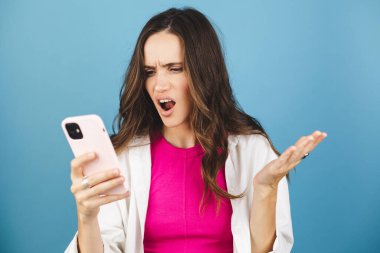 Mad ve şok genç esmer güzel kadın mavi arka planda izole cep telefonu kullanıyor. Bağıran mutsuz kadın cep telefonunu tutuyor. Hayal kırıklığına uğramış üzgün bayan, haberleri dehşete düşürdü. Bu da ne? Olamaz..