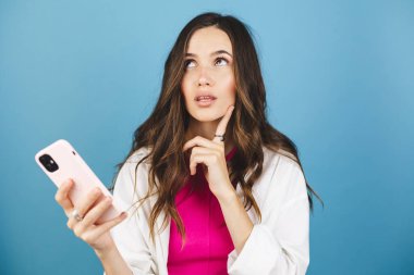 Esmer, kıvırcık kadın soruya nasıl cevap verileceğini düşünür, bir şey hakkında derin düşünür, modern cep telefonu kullanır, iyi bir mesaj uydurmaya çalışır, işaret parmağını tutar, mavi arka planda tek başına durur..