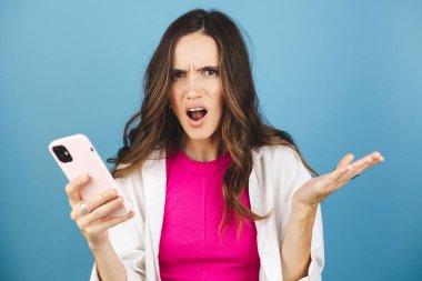 Mad ve şok genç esmer güzel kadın mavi arka planda izole cep telefonu kullanıyor. Bağıran mutsuz kadın cep telefonunu tutuyor. Hayal kırıklığına uğramış üzgün bayan, haberleri dehşete düşürdü. Bu da ne? Olamaz..