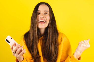 Eğlenceli genç kadın cep telefonu kullanıyor ve sarı arka plan stüdyosunda izole edilmiş yumruklarını sallıyor. Kız internet üzerinden indirimli hediye kuponu kazanan akıllı telefon uygulamaları kullanıyor..