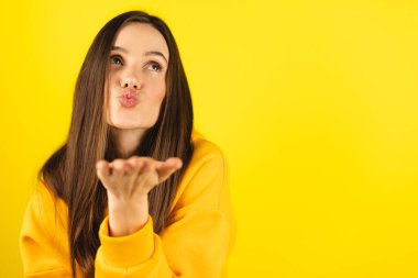 Sarı kapüşonlu giyen mutlu bir kadın sarı stüdyoya hava öpücüğü gönderir. Çekici kız öpücük yolluyor. Kadın özgürlüğü yaşam tarzı. Çekici şıklığın kameraya öpücüğü göndermesi. Kadın aşk hikayesi. Duygu..