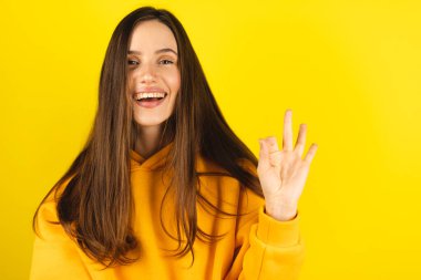 Mutlu, genç ve çekici bir kadın parmaklarıyla işaret veriyor. Her şeyin süper, neşe dolu olduğunu söylemeye çalışıyor. Sarı arka planda izole edilmiş kameraya bakıyor..