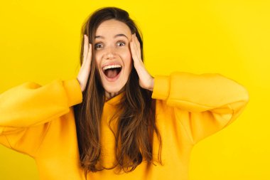 İyimser kadın reaksiyon elleri başını tutuyor wow, birinden müthiş bir hediye almaktan mutlu, yüksek sesle bağırıyor, sarı kapüşonlu giyinmiş, sarı arka planda soyutlanmış. Heyecanlı kadın çığlıkları, mutlu çığlıklar..