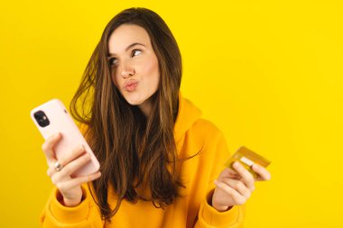 İnternetten alışveriş. Gülümseyen esmer kadının portresi akıllı telefon uygulamasında plastik kredi kartıyla ödeme yapması, cep telefonu ve banka kartıyla sarı arka planda durması. Mutlu duygular.