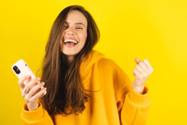 Fun young woman hold in hand use mobile cell phone doing winner gesture clenching fists isolated on yellow background studio. Girl use smartphone apps winning online celebrating discount gift voucher. clipart