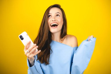 Eğlenceli genç kadın cep telefonu kullanıyor ve sarı arka planda izole edilmiş yumruklarını sallıyor. Akıllı telefon uygulaması kullanan kız internette indirim kuponu kazanıyor..