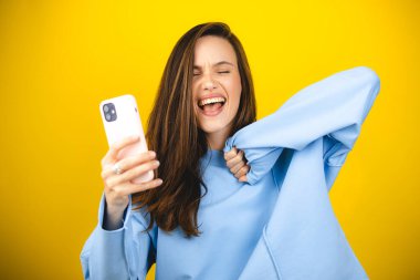 Fun young woman hold in hand use mobile cell phone doing winner gesture clenching fists isolated on yellow background. Girl using smartphone apps winning online celebrating discount gift voucher. clipart