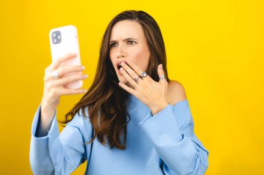 Sarı arka planda izole edilmiş cep telefonu kullanan genç ve güzel bir kadın. Bağıran mutsuz kadın cep telefonunu tutuyor. Hayal kırıklığına uğramış üzgün bayan, haberleri dehşete düşürdü. Bu da ne? Olamaz.