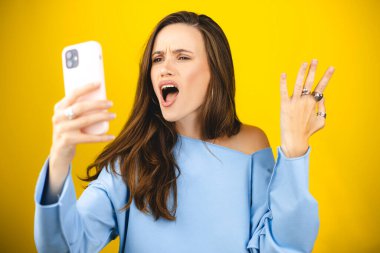 Sarı arka planda izole edilmiş cep telefonu kullanan genç ve güzel bir kadın. Bağıran mutsuz kadın cep telefonunu tutuyor. Hayal kırıklığına uğramış üzgün bayan, haberleri dehşete düşürdü. Bu da ne? Olamaz.