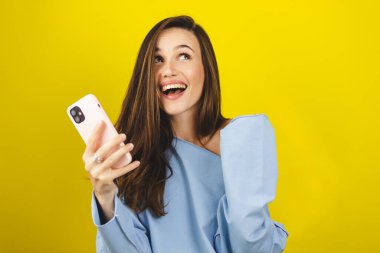 Fun young woman hold in hand use mobile cell phone doing winner gesture clenching fists isolated on yellow background. Girl using smartphone apps winning online celebrating discount gift voucher. clipart