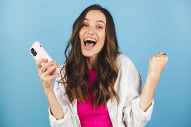 Eğlenceli genç kadın cep telefonu kullanıyor. Mavi arka plan stüdyosunda izole edilmiş yumruklarını sallıyor. Akıllı telefon uygulaması kullanan kız internette indirim kuponu kazanıyor.