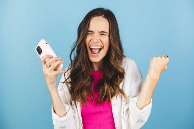 Fun young woman hold in hand use mobile cell phone doing winner gesture clenching fists isolated on blue background studio. Girl using smartphone apps winning online celebrating discount gift voucher clipart