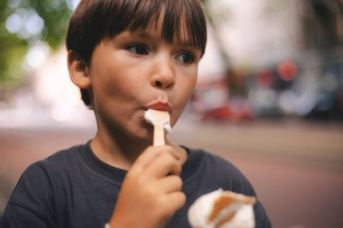 Küçük esmer çocuk gözlemeli mısır ve kaşıkla dondurma yiyor. Mutlu bir çocukluk. Çocuk şehir caddesinde yürür.. 
