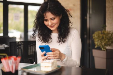 Telefon kullanan kıvırcık saçlı genç bir kadın kafeteryada oturuyor. Akıllı telefon, mesajlar, telefon görüşmeleri, mektuplar, mesajlar, mesajlar, mesajlar, sohbet ya da mesajlar. Kız mesajlaşması, ödemeli telefon, mutlu..