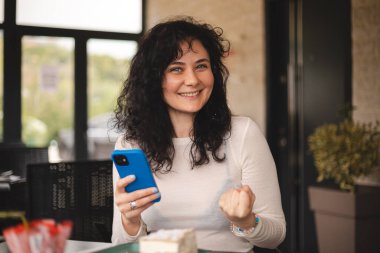 Neşeli kıvırcık esmer kadın cep telefonuyla video oyunu oynuyor, iyi haberler okuyor, piyangoyu kazanmayı kutluyor, kazanan hareketi yaparken çok mutlu ve neşeli hissediyor, masada oturuyor..