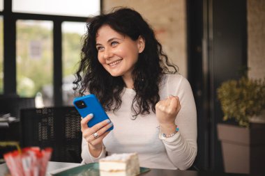 Neşeli kıvırcık esmer kadın cep telefonuyla video oyunu oynuyor, iyi haberler okuyor, piyangoyu kazanmayı kutluyor, kazanan hareketi yaparken çok mutlu ve neşeli hissediyor, masada oturuyor..