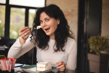 Genç kıvırcık esmer kadın kremalı pasta yiyor, çatalla kafede oturuyor. Yemek günü konseptini aldatmak. Kadın ekkin yemek için iştahla hazırlanıyor. Restorandaki pastanenin tadını çıkar. Vay canına..