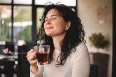 Kahvede oturan mutlu esmer, kıvırcık kadın, biriyle güzel bir sohbet ettiğinde, çayın tadını ve kokusunu alır. Beyaz giysili kadın çay içiyor, gülümsüyor.. 