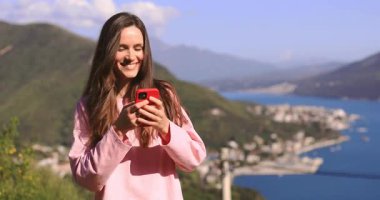 Harika uygulama ya da web sitesi. Heyecanlı gülümseyen kadın cep telefonunu kullanıyor, açık havada dağ manzarasına karşı duruyor. Elinde alet tutan olumlu bir kadın, mesaj, mesaj, kameraya yan yana bak..