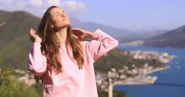 Mutlu esmer, uzun saçlı kadın, dağ ve denizin karşısında duruyor kablosuz kulaklıklarla müzik dinliyor, saç yapıyor, müzik dinliyor pembe uzun kollu elbiseler giyiyor..