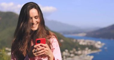 Harika uygulama ya da web sitesi. Heyecanlı gülümseyen kadın cep telefonunu kullanıyor, açık havada dağ manzarasına karşı duruyor. Elinde alet tutan olumlu kadın, mesaj, mesaj, kameraya bak..