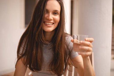 Mutlu çekici kadın evin yanında otururken bir bardak su içiyor. Genç, güzel bir kız uyandıktan sonra bardakta temiz mineral tutuyor ve sabah sağlık hizmeti için yudumluyor. Sıvı kaybı.