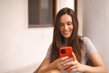 Cep telefonu tutan ve evin yanında otururken akıllı telefona bakan genç ve güzel bir kadın. Mutlu kız cep telefonu kullanıyor ve flört ediyor. Gülümseyen kadın telefonu tut, iyi haberler oku, mesaj bırak..