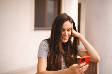 Cep telefonu tutan ve evin yanında otururken akıllı telefona bakan genç ve güzel bir kadın. Mutlu kız cep telefonu kullanıyor ve flört ediyor. Gülümseyen kadın telefonu tut, iyi haberler oku, mesaj bırak..