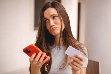 Kumral, uzun saçlı, kısa saçlı bir kadın kredi kartı kullanıyor. Akıllı telefon, e-ödemeyle ilgili sorunlar yaşıyor. Parasızlık, ödeme güvensizliği, yetersiz fonlar yüzünden kendini rahatsız hissediyor. Dolandırıcılık mağduru..