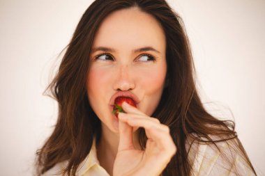 Kapatın esmer yüzlü güzel, hoş, hoş, lüks kadın portresi çekici, dalgalı saçlı kadın ısırıyor, tadıyor, çilek yiyor, neşeli görünüyor, tişört giyiyor.. 