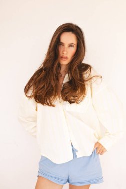 Uzun kahverengi saçlı, açık renk çizgili tişörtlü ve mavi şortlu, rahat ve rahat bir şekilde içeride poz veren genç bir kadın. Doğal güzellik, minimalist moda ve rahat ev atmosferi..