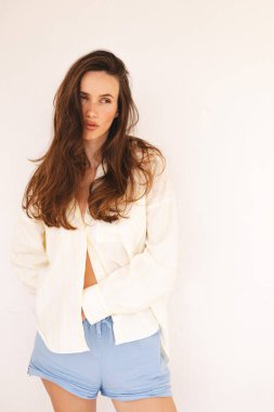 Uzun kahverengi saçlı, açık renk çizgili tişörtlü ve mavi şortlu, rahat ve rahat bir şekilde içeride poz veren genç bir kadın. Doğal güzellik, minimalist moda ve rahat ev atmosferi..