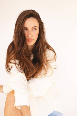 Uzun kahverengi saçlı, açık renk çizgili tişörtlü ve mavi şortlu, rahat ve rahat bir şekilde içeride poz veren genç bir kadın. Doğal güzellik, minimalist moda ve rahat ev atmosferi..