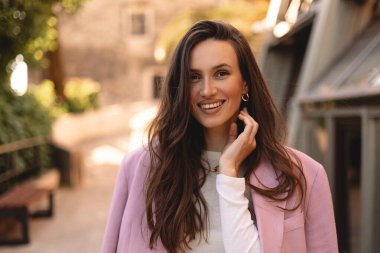 Açık havada gülümseyen uzun saçlı, pembe ceketli ve beyaz tişörtlü çekici bir genç kadın. Şehir yaşam tarzı, doğal güzellik, günışığında moda portresi. El ele tutuşun..