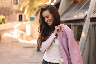 Genç bir kadın gülümsüyor ve aşağıya bakıyor pembe ceketini dışarı yerleştirirken, doğal ışığa güveni ve kendine güveni olan, samimi moda portresi