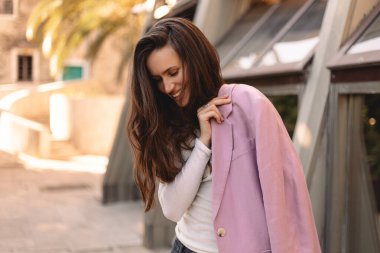 Genç bir kadın gülümsüyor ve aşağıya bakıyor pembe ceketini dışarı yerleştirirken, doğal ışığa güveni ve kendine güveni olan, samimi moda portresi