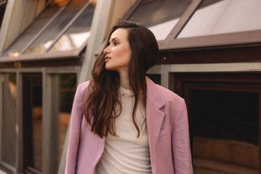 Esmer, uzun dalgalı saçlı, pembe ceketli, ofis binasının yanında duran ve yan tarafa bakan. Zarif, şehvetli bir patron, düşünceli görünüyor, yandan bak..