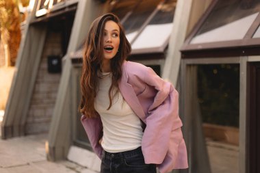 Pembe bir ceketle kendinden emin poz veren enerjik genç bir kadın şehir ortamında neşe ve tarz yayıyor. O hareket ederken dalgalı saçlar uçuşuyor. Moda ve yaşam tarzı için coşkulu ve tasasız bir anı yakalıyor.. 