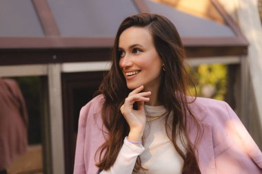 Düşünceli, uzun dalga saçlı, pembe ceketli, zarif bir jest ve sakin bir ifadeyle dışarıda poz veren genç ve güzel şeyler düşünen bir kız..