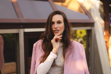 Düşünceli, uzun dalga saçlı, pembe ceketli, zarif bir jest ve sakin bir ifadeyle dışarıda poz veren genç ve güzel şeyler düşünen bir kız..