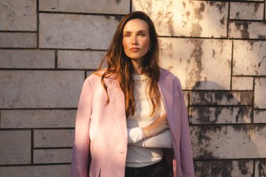  Pembe ceketli, modern taş bir duvara karşı çapraz kollarla poz veren kendinden emin genç bir kadın, sıcak güneş ışığında duruyor. Doğal hafif moda portresi, şık bir duruşu ve rahat duruşu var..