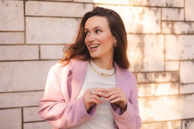 Ofis binasının arka planında duran kadın girişimci yan tarafa bakıyor. Başarılı gülümseyen, beyaz takım elbiseli, omuzlarında pembe ceketi olan bir kadın. Çapraz kollu kadın işçi mutlu görünüyor.