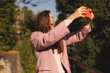 Pembe ceketli, uzun kahverengi saçlı mutlu genç bir kadın kırmızı akıllı telefonuyla selfie çekiyor. Güneşli bir havada balkonda dikilirken gülümsüyor ve gülüyor. Etrafı yeşil ağaçlar ve gökyüzüyle çevrili..