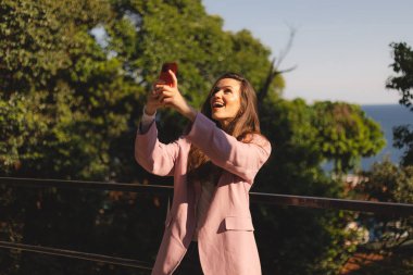 Pembe ceketli, uzun kahverengi saçlı mutlu genç bir kadın kırmızı akıllı telefonuyla selfie çekiyor. Güneşli bir havada balkonda dikilirken gülümsüyor ve gülüyor. Etrafı yeşil ağaçlar ve gökyüzüyle çevrili..