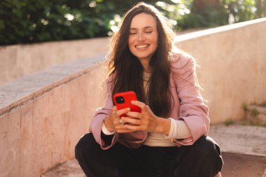 Pembe ceketli gülümseyen genç kadın taş basamaklarda oturuyor, akıllı telefon kullanıyor, mesajlaşıyor ve gülüyor. Rahat duruş, bacak bacak üstüne atma, güneşli günün tadını çıkarma..