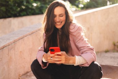 Pembe ceketli gülümseyen genç kadın taş basamaklarda oturuyor, akıllı telefon kullanıyor, mesajlaşıyor ve gülüyor. Rahat duruş, bacak bacak üstüne atma, güneşli günün tadını çıkarma..