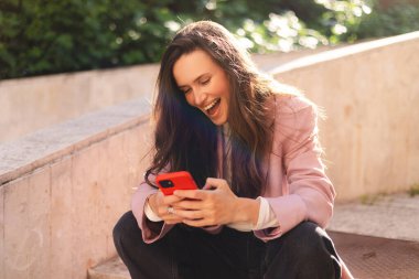 Keyifli esmer kız, şaşırtıcı duygular sergiliyor ve cep telefonunu sokakta merdivenlerde otururken kullanıyor. Cep telefonuyla internette sörf yapan, pembe ceket giyen sürpriz kız..